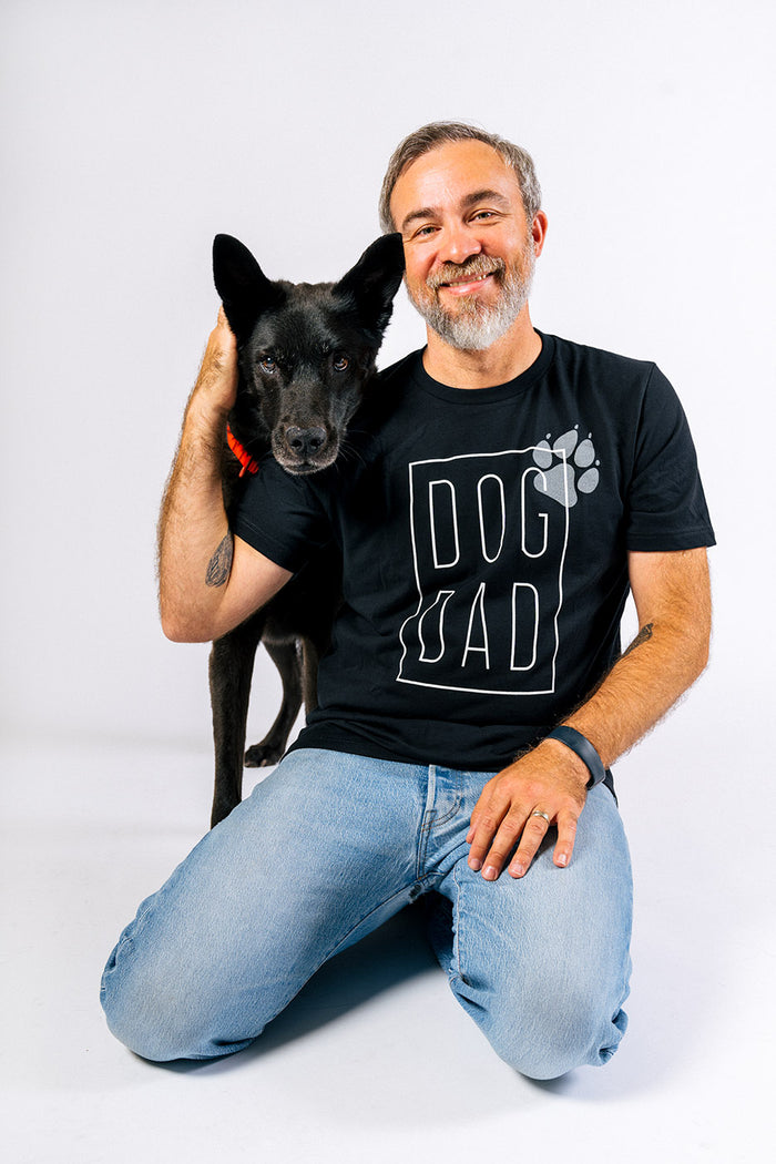 P.L.A.Y. Dog Dad Humanitee - T-Shirt modeled on dog dad sitting on his knees with his beautiful black dog over his shoulder both looking at the camera smiling