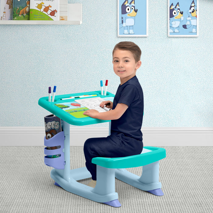 Child sitting at a adjustable desk with a blue and green color scheme, smiling in a room with light blue walls and cartoon character posters.