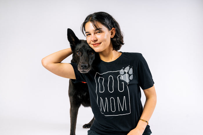 P.L.A.Y. Dog Mom Humanitee - T-Shirt modeled on woman sitting with beautiful black dog putting her head on her shoulder, both smiling and looking at the camera