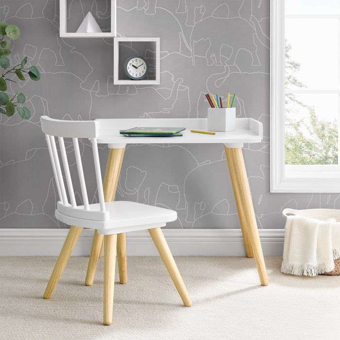 A child sitting at a white Essex desk with a beige chair, which has natural-colored tapered legs. The desk is against a wall with wallpaper, and there is a plant and a piece of furniture in the background.