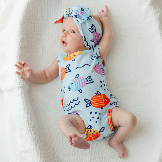A baby lying on a white textured blanket, wearing a light blue one-piece swimsuit and matching hat with colorful fish and wave patterns.