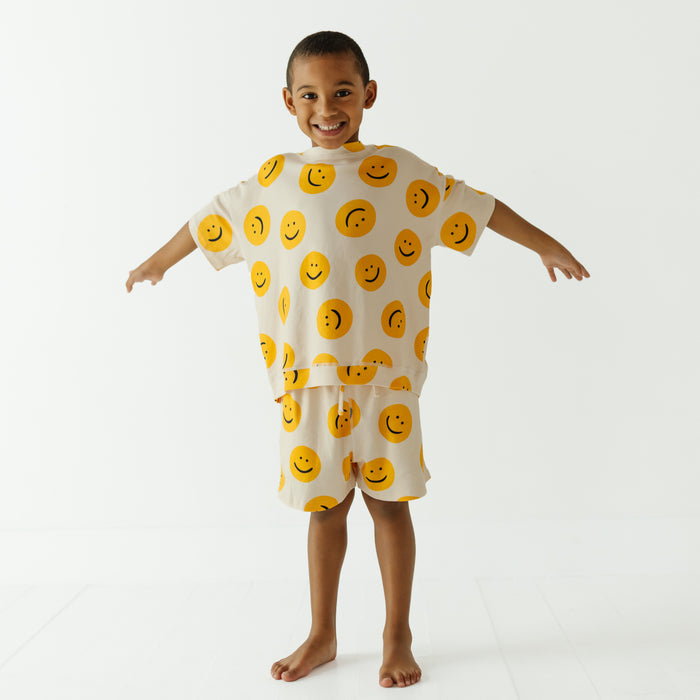 A smiling young boy stands barefoot against a clean white studio background, wearing a cream-colored pajama set with a repeating yellow smiley face pattern.
