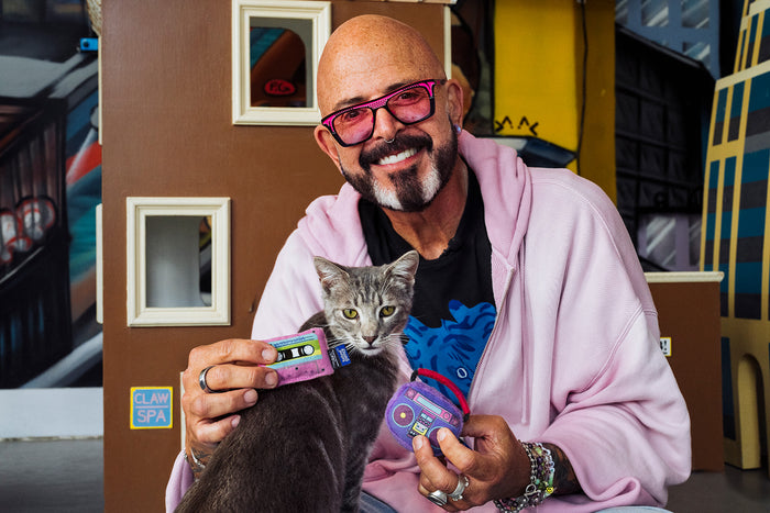 Jackson Galaxy holding a gray tabby cat and the Jackson Galaxy x Feline Frenzy Playback Duo in his hands in front of a cardboard house.