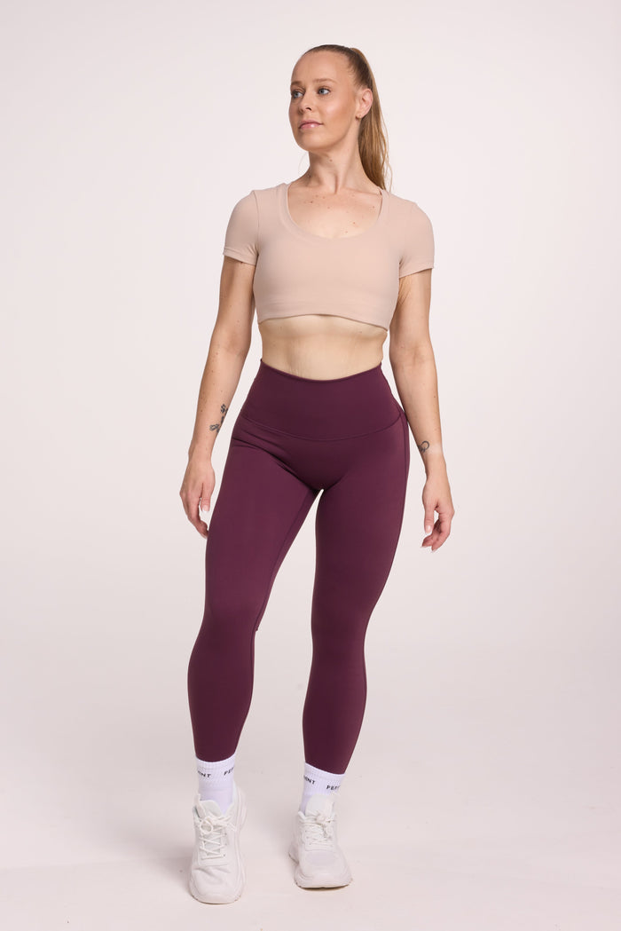 Buttery soft high waisted women's gym tights with curved back seam stitched detailing for a butt shaping effect, and no front rise seam, in plum purple paired with an V neck women's crop t-shirt with a supportive elasticated hem and triple layered fabric with removable pads for extra support in beige