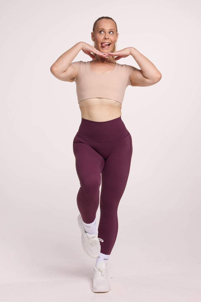 Buttery soft high waisted women's gym tights with curved back seam stitched detailing for a butt shaping effect, and no front rise seam, in plum purple paired with an V neck women's crop t-shirt with a supportive elasticated hem and triple layered fabric with removable pads for extra support in beige