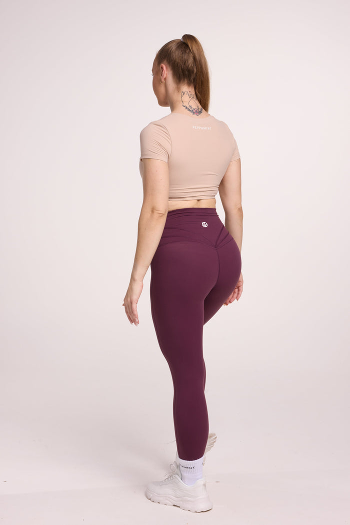 Buttery soft high waisted women's gym tights with curved back seam stitched detailing for a butt shaping effect, and no front rise seam, in plum purple paired with an V neck women's crop t-shirt with a supportive elasticated hem and triple layered fabric with removable pads for extra support in beige