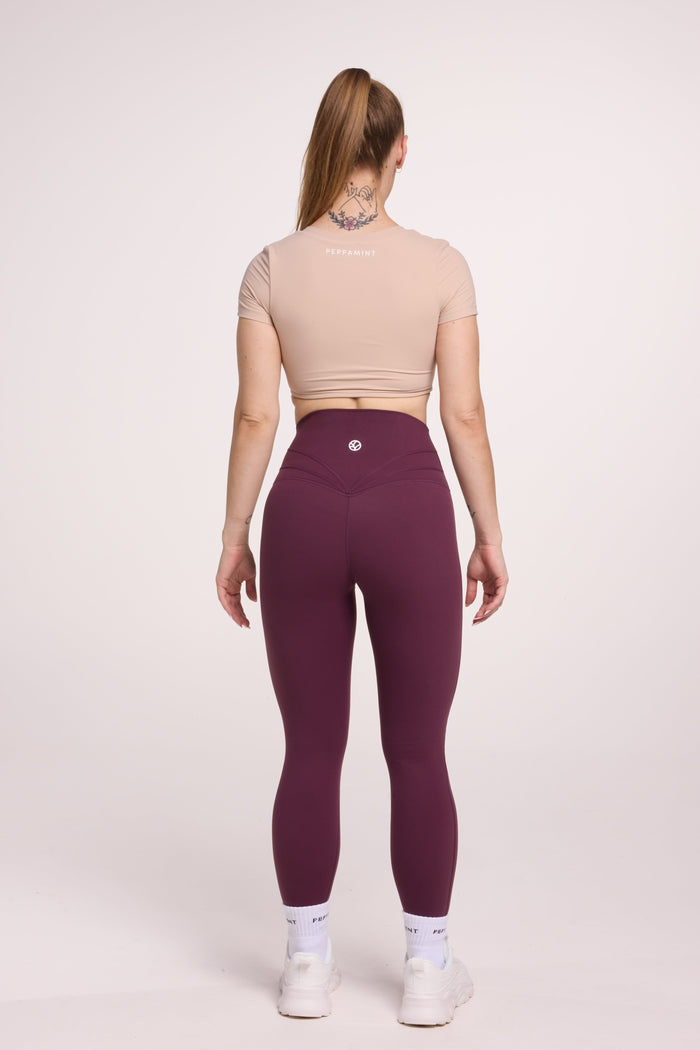 Buttery soft high waisted women's gym tights with curved back seam stitched detailing for a butt shaping effect, and no front rise seam, in plum purple paired with an V neck women's crop t-shirt with a supportive elasticated hem and triple layered fabric with removable pads for extra support in beige