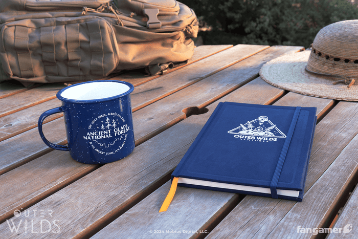 A photograph of two new Outer Wilds items on a weathered wooden table, with a camping pack and a hat at the edge of the frame. A navy Outer Wilds camping mug says "Ancient Glade National Forest," with a speckled finish and a white interior. The navy Hatchling's Training Log journal has the Outer Wilds Ventures logo stamped in foil across its cover, with an orange ribbon bookmark and an elastic enclosure.