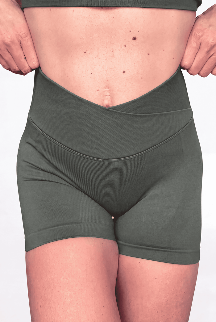 A close-up of a person wearing high-waisted athletic shorts with V crossover waistband in a sage green colour, holding the sides of the waistband to show its stretch and shape.