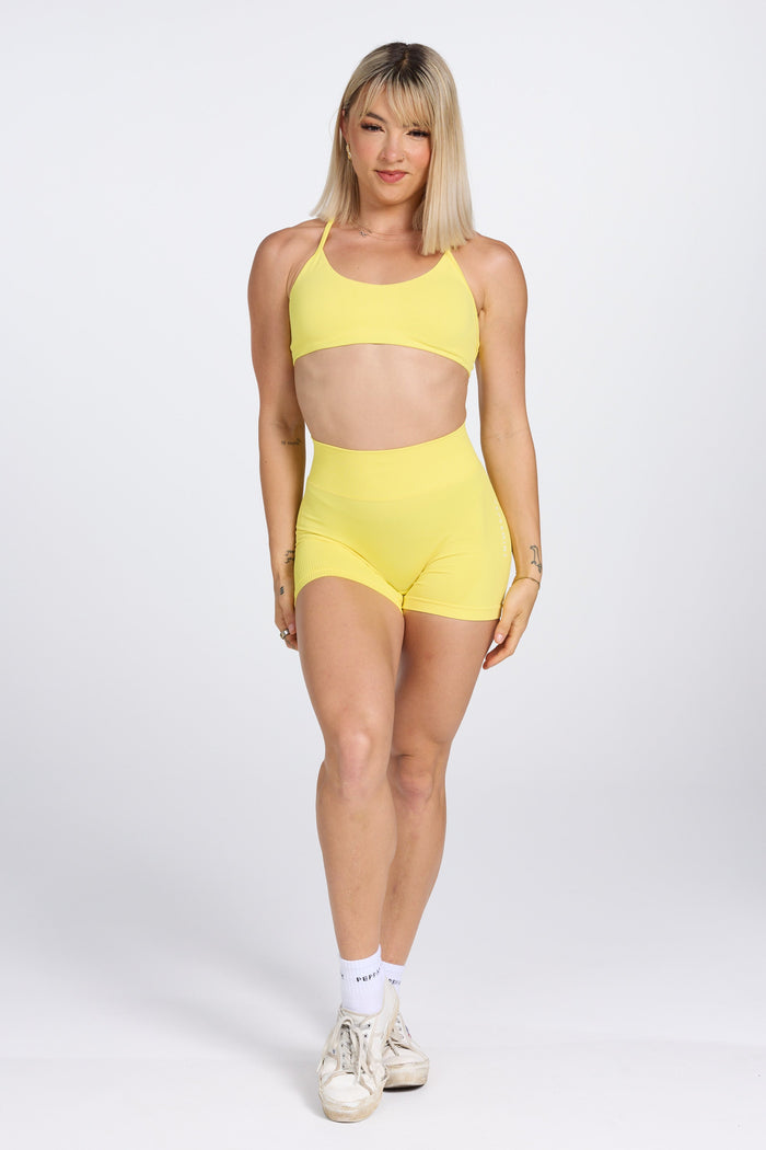 Seamless women’s gym bra with an ultra thin racerback strap design, with white PEPPAMINT branding on the back strap paired with high waisted gym shorts with booty contouring panels, invisible scrunch technology, sweetheart shaped back seams and thick supportive fabric in buttercup yellow