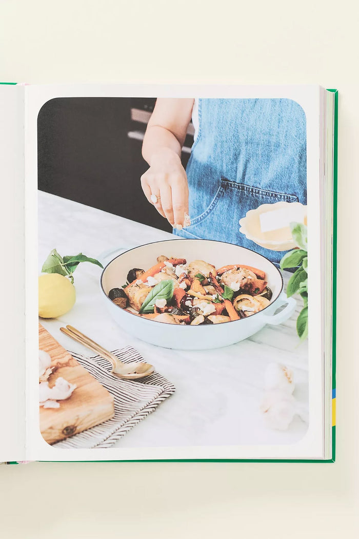 A hardcover cookbook or recipe book shown open on a light cream background. The cover features a large photograph of a person in a blue apron seasoning a bowl of pasta on a kitchen counter, with a lemon and wooden spoon nearby.